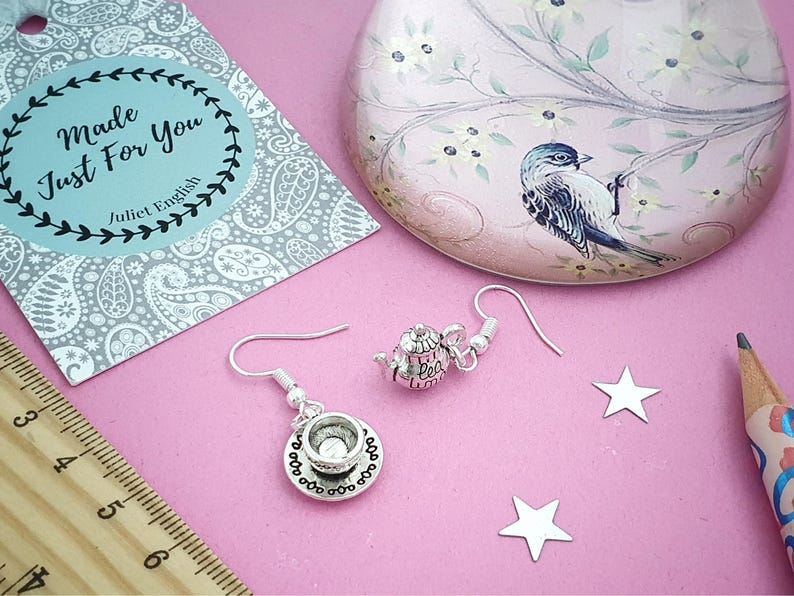 May include: Silver-toned teapot and teacup earrings with a matching design. The earrings are displayed on a pink surface with star-shaped confetti, a decorative tin, and a tag that reads "Made Just For You Juliet English."