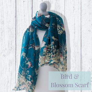 May include: A teal blue scarf with a white and pink floral pattern and small birds. The scarf has a gradient effect with the floral pattern fading out at the bottom. The text "Bird & Blossom Scarf" is printed on a light blue background below the scarf.