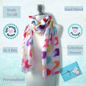 May include: A white scarf with a colorful print of cartoon birds. The scarf is displayed on a mannequin and is ready to be gifted. The image also shows a blue gift box with a ribbon and a gift tag.