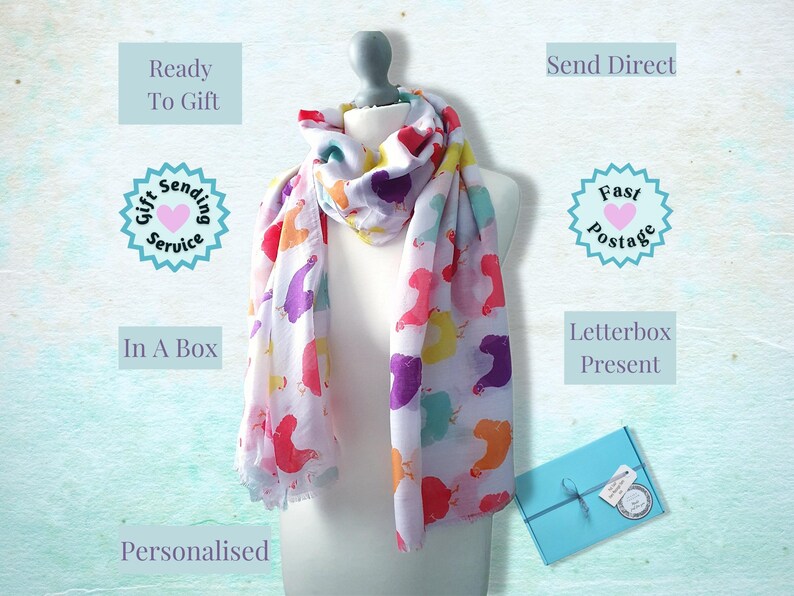 May include: A white scarf with a colorful print of cartoon birds. The scarf is displayed on a mannequin and is ready to be gifted. The image also shows a blue gift box with a ribbon and a gift tag.