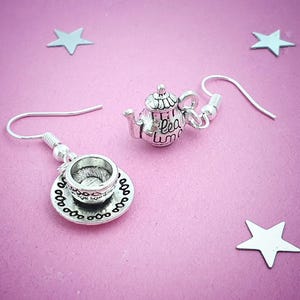 May include: Silver-toned teapot and teacup earrings with a delicate design. The teapot has the words "tea time" on it. The earrings are on a pink background with silver star-shaped decorations.