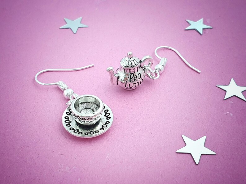 May include: Silver-toned teapot and teacup earrings with a delicate design. The teapot has the words "tea time" on it. The earrings are on a pink background with silver star-shaped decorations.