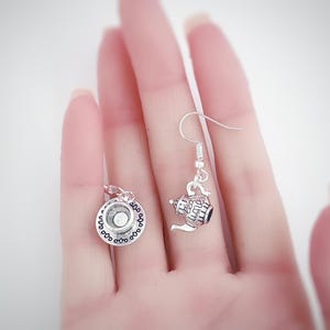 May include: Silver-toned teapot and teacup earrings. The earrings feature a detailed teapot charm with a handle and spout, and a teacup charm with a saucer. The charms are suspended from silver-colored ear wires. The earrings are a charming accessory.