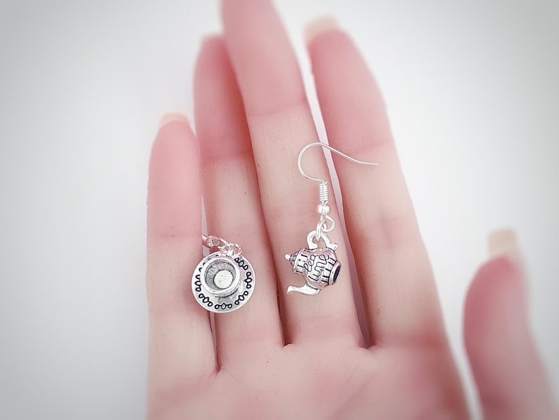 May include: Silver-toned teapot and teacup earrings. The earrings feature a detailed teapot charm with a handle and spout, and a teacup charm with a saucer. The charms are suspended from silver-colored ear wires. The earrings are a charming accessory.
