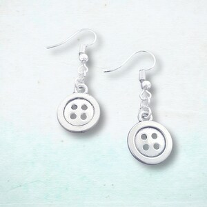 May include: A pair of silver button earrings. Each earring has a button charm with four holes, hanging from a simple hook. The earrings are set against a light, textured background.