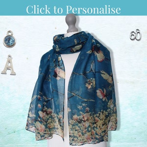 May include: A teal blue scarf with a floral and bird print. The scarf is displayed on a mannequin. There are three silver charms: a letter A, a number 60, and a blue gemstone.
