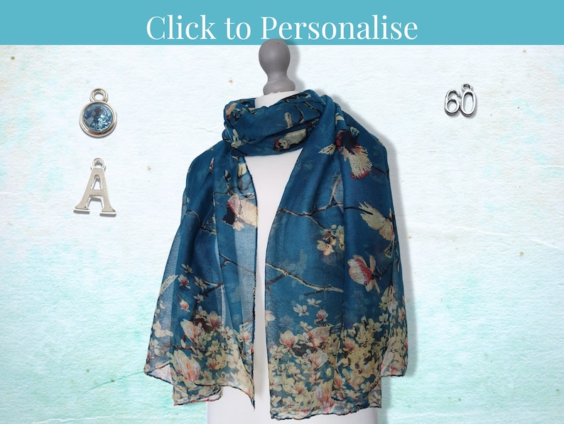 May include: A teal blue scarf with a floral and bird print. The scarf is displayed on a mannequin. There are three silver charms: a letter A, a number 60, and a blue gemstone.