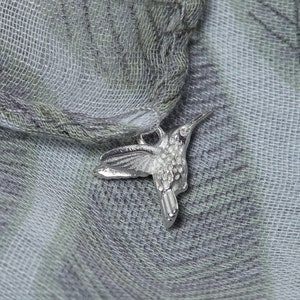 May include: A silver hummingbird charm with a detailed design, hanging from a light gray and white patterned scarf.