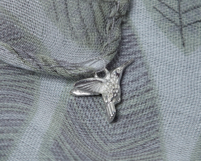 May include: A silver hummingbird charm with a detailed design, hanging from a light gray and white patterned scarf.