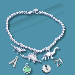 May include: Silver beaded bracelet featuring dinosaur charms, a letter "A" charm, a green gemstone charm, and a "21" charm. The bracelet has a lobster clasp. The background is teal.
