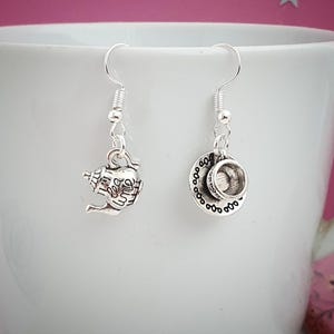 May include: Silver-toned dangle earrings featuring a teapot and teacup design. The teapot earring has a handle and spout, while the teacup earring includes a saucer. Both earrings are suspended from a silver fish hook.
