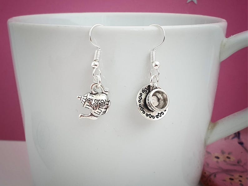 May include: Silver-toned dangle earrings featuring a teapot and teacup design. The teapot earring has a handle and spout, while the teacup earring includes a saucer. Both earrings are suspended from a silver fish hook.