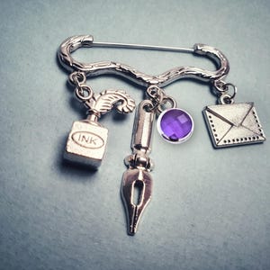 May include: A silver-toned safety pin brooch featuring three dangling charms: an inkwell with a quill pen, a fountain pen nib, and a sealed envelope. A small, round, faceted purple gemstone is attached to the centre of the brooch assembly.