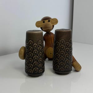 Może przedstawiać: Dwie brązowe solniczki i pieprzniczki ceramiczne z kwiatowym wzorem i drewnianymi uchwytami. Drewniana figurka małpy jest umieszczona pomiędzy solniczkami i pieprzniczkami. Przedmioty znajdują się na białej powierzchni.