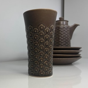 Può includere: Un bicchiere in ceramica marrone con un motivo floreale, una teiera e dei piatti. Il bicchiere è il punto focale, con un motivo floreale in rilievo ripetuto. La teiera e i piatti sono sullo sfondo, creando un set coeso. Gli oggetti sono su una superficie bianca.