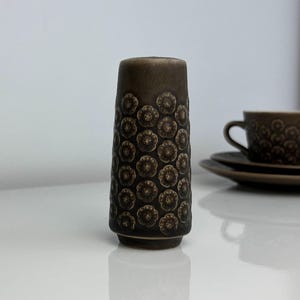 Può includere: Un vaso in ceramica marrone con un motivo floreale, accanto a una tazza e un piattino abbinati. Il vaso è cilindrico, alto circa 15 cm, e la tazza e il piattino sono anch'essi marroni. Gli oggetti sono su una superficie bianca.