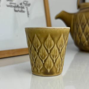 Puede incluir: Una pequeña taza de cerámica verde oliva con un diseño de hojas. La taza está junto a una tetera a juego. La taza tiene un borde blanco. El fondo incluye un mapa enmarcado y una superficie blanca. La taza mide unos 7 cm de alto.