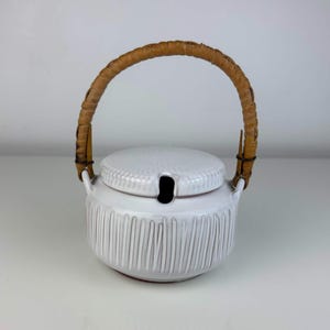 May include: A white ceramic lidded jar with a textured vertical ribbed pattern on the body and a matching lid. The jar features a curved, light brown woven rattan handle attached to two side loops and a small notch in the lid for a spoon.
