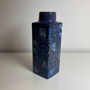 May include: A rectangular, dark blue ceramic vase with a square base and a slightly narrower neck. The vase features a repeating geometric pattern in shades of blue and black. The vase is approximately 8 inches tall.