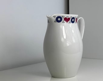 May include: White ceramic pitcher with a decorative rim featuring blue circles and red hearts. The pitcher has a curved handle and a glossy finish, suitable for serving beverages.