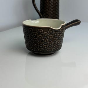 Può includere: Una salsiera in ceramica marrone con manico e motivo floreale. L'interno è color crema chiaro. Sullo sfondo è visibile una brocca abbinata. La salsiera è un oggetto decorativo per la cucina.