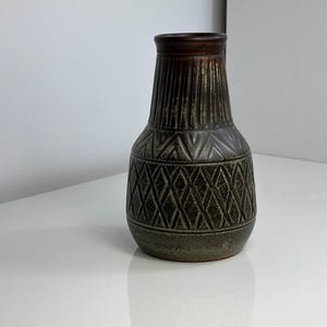 Puede incluir: Un jarrón decorativo de cerámica con borde marrón oscuro y cuerpo texturizado y estampado. El jarrón presenta un diseño de diamantes y líneas verticales en tonos verdes y marrones. El jarrón mide aproximadamente 20 cm de altura.