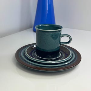 Puede incluir: Una taza de cerámica color turquesa con asa se encuentra sobre un juego de platos a juego. Los platos tienen anillos concéntricos de color, incluyendo turquesa, marrón y azul marino. Un jarrón alto y azul está en el fondo.