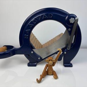 Puede incluir: Una rebanadora de pan azul marino con la palabra "RAADVAD" en el arco superior. La rebanadora tiene un mango de madera y una cuchilla de acero inoxidable. Un pequeño mono de madera está delante de la rebanadora.