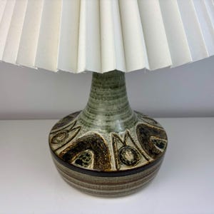 May include: A ceramic table lamp with a cream-coloured pleated lampshade. The base is a mix of green, brown, and tan with a textured, patterned design. The lamp has a bulbous shape and a narrow neck.