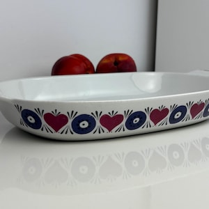 Puede incluir: Fuente de horno ovalada de cerámica blanca con un borde decorativo de corazones rojos y círculos azules alternados. La fuente tiene asas pequeñas en cada extremo y está sobre una superficie blanca. Dos melocotones rojos están en el fondo.