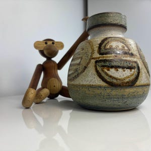 Puede incluir: Un jarrón de cerámica con superficie texturizada y diseños abstractos en marrón y beige. Una figura de mono de madera con extremidades móviles está sentada junto al jarrón. El jarrón tiene borde y base verdes.