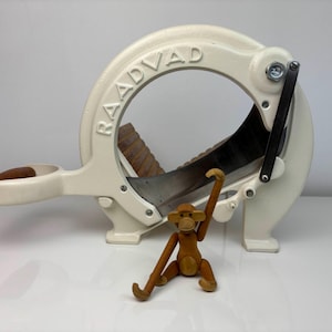 Puede incluir: Cortadora de pan RAADVAD de color crema con una cuchilla curva y un mango. Un pequeño juguete de mono de madera está colocado delante de la cortadora. La cortadora tiene la palabra RAADVAD grabada en el lateral.