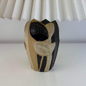 May include: A decorative table lamp with a cream-colored pleated lampshade. The lamp base is a vase shape, featuring a black and tan floral design. The lamp is a unique home decor item.