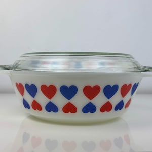 Può includere: Pirofila bianca con coperchio in vetro trasparente. Il piatto è decorato con cuori rossi e blu alternati. Il piatto ha due manici ed è appoggiato su una superficie bianca.