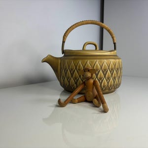 以下が含まれることがあります： 編み込みの取っ手と葉の模様が付いた、オリーブグリーンのセラミックティーポット。木製の猿の置物がティーポットの前に置かれています。 ティーポットには注ぎ口と小さなハンドルが付いた蓋があります。