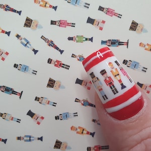 May include: A red and white striped nail wrap with a design of four nutcracker soldiers. The soldiers are wearing traditional costumes with red, blue, and gold accents.