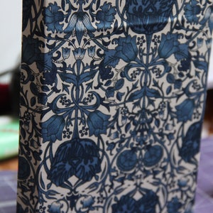 May include: A hardback book featuring a fabric cover with a repeating pattern of dark blue flowers, leaves, and stems on a cream-coloured background. The book is standing upright, showing the front cover and the spine with white pages visible at the top.