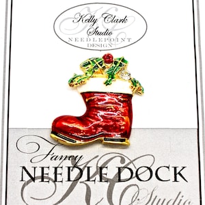 Puede incluir: Un pin de media navideña de color rojo y dorado con un puño blanco y acebo verde con una baya roja en la parte superior. El pin está en una tarjeta blanca con el texto "Kelly Clark Studio Needlepoint Design" y "Fancy Needle Dock KC Studio".
