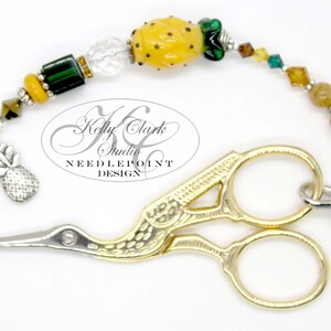 May include: Gold-toned bird-shaped embroidery scissors with a pineapple charm and a beaded chain. The chain features green, yellow, and brown beads. The text "Kelly Clark Studio NEEDLEPOINT DESIGN" is visible in the background.