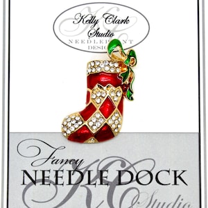 May include: A red and gold Christmas stocking pin with rhinestones and a green bow. The pin is on a white card with the text "Kelly Clark Studio Needlepoint Design" and "Fancy Needle Dock KC Studio".