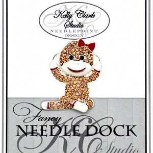 May include: A brown and white monkey with a red bow, covered in sparkly rhinestones, is sitting with its hands covering its ears. The monkey is on a white card with the text "Kelly Clark Studio Needlepoint Design" and "Fancy Needle Dock KC Studio".