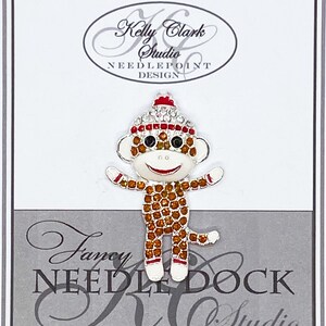 May include: A close-up of a small, sparkly monkey wearing a red and white knit hat. The monkey is made of rhinestones and has a white face with red lips and black eyes. The monkey is standing on a white background with the text "Kelly Clark Studio Needlepoint Design" and "Fancy Needle Dock KC Studio" printed on the background.