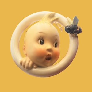 May include: A vintage ceramic figurine featuring a surprised baby face with rosy cheeks, surrounded by a white ring. A black and yellow bee is perched on the ring. The figurine has a yellow background.