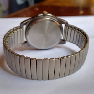 K&ouml;nnte beinhalten: Eine silberfarbene Armbanduhr mit einem dehnbaren Armband. Das Zifferblatt der Uhr ist nicht sichtbar, aber das Geh&auml;use auf der R&uuml;ckseite ist hellsilberfarben. Das Armband besteht aus kleinen, rechteckigen Metallgliedern, die sich dehnen.