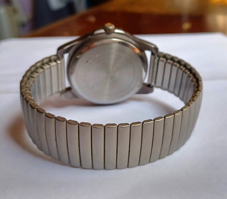 K&ouml;nnte beinhalten: Eine silberfarbene Armbanduhr mit einem dehnbaren Armband. Das Zifferblatt der Uhr ist nicht sichtbar, aber das Geh&auml;use auf der R&uuml;ckseite ist hellsilberfarben. Das Armband besteht aus kleinen, rechteckigen Metallgliedern, die sich dehnen.