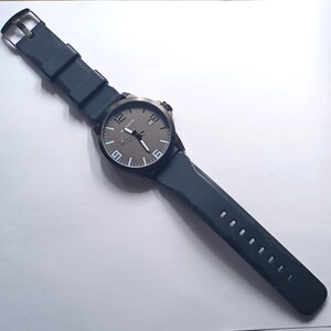 K&ouml;nnte beinhalten: Eine Armbanduhr mit dunkelgrauem Zifferblatt, wei&szlig;en Ziffern auf den 12-, 6- und 9-Uhr-Positionen, einem Datumsfenster auf 3 Uhr und einem schwarzen Silikonarmband mit Metallschnalle. Das Uhrengeh&auml;use ist schwarz und hat eine passende Krone.