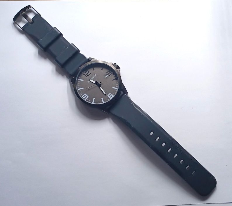 K&ouml;nnte beinhalten: Eine Armbanduhr mit dunkelgrauem Zifferblatt, wei&szlig;en Ziffern auf den 12-, 6- und 9-Uhr-Positionen, einem Datumsfenster auf 3 Uhr und einem schwarzen Silikonarmband mit Metallschnalle. Das Uhrengeh&auml;use ist schwarz und hat eine passende Krone.