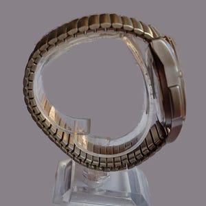 K&ouml;nnte beinhalten: Eine silberfarbene Armbanduhr mit rechteckigem Zifferblatt und einem flexiblen, dehnbaren Metallarmband. Die Uhr wird auf einem transparenten Kunststoffst&auml;nder pr&auml;sentiert, der das Design des Armbands hervorhebt. Das Zifferblatt der Uhr ist silberfarben mit dunkler Anzeige.