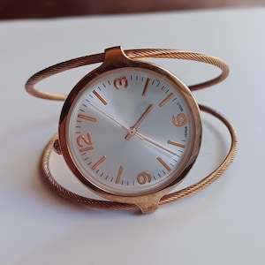 May include: Rose gold-toned cuff watch with a round face. The watch features a silver-colored dial with rose gold-toned numerals and hands. The cuff is made of twisted metal and has a minimalist design.