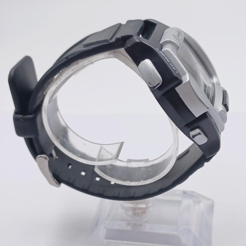 May include: Black and silver digital watch with a rectangular face and a black rubber strap. The watch features silver buttons and a clear display. The watch is displayed on a clear plastic stand.